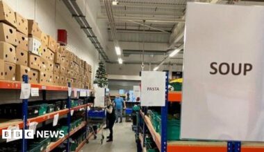 A long aisle in a warehouse stacked with boxes and cans of food. There are signs saying "Pasta" and "Soup". A woman with a trolley is in the distance picking up cans.