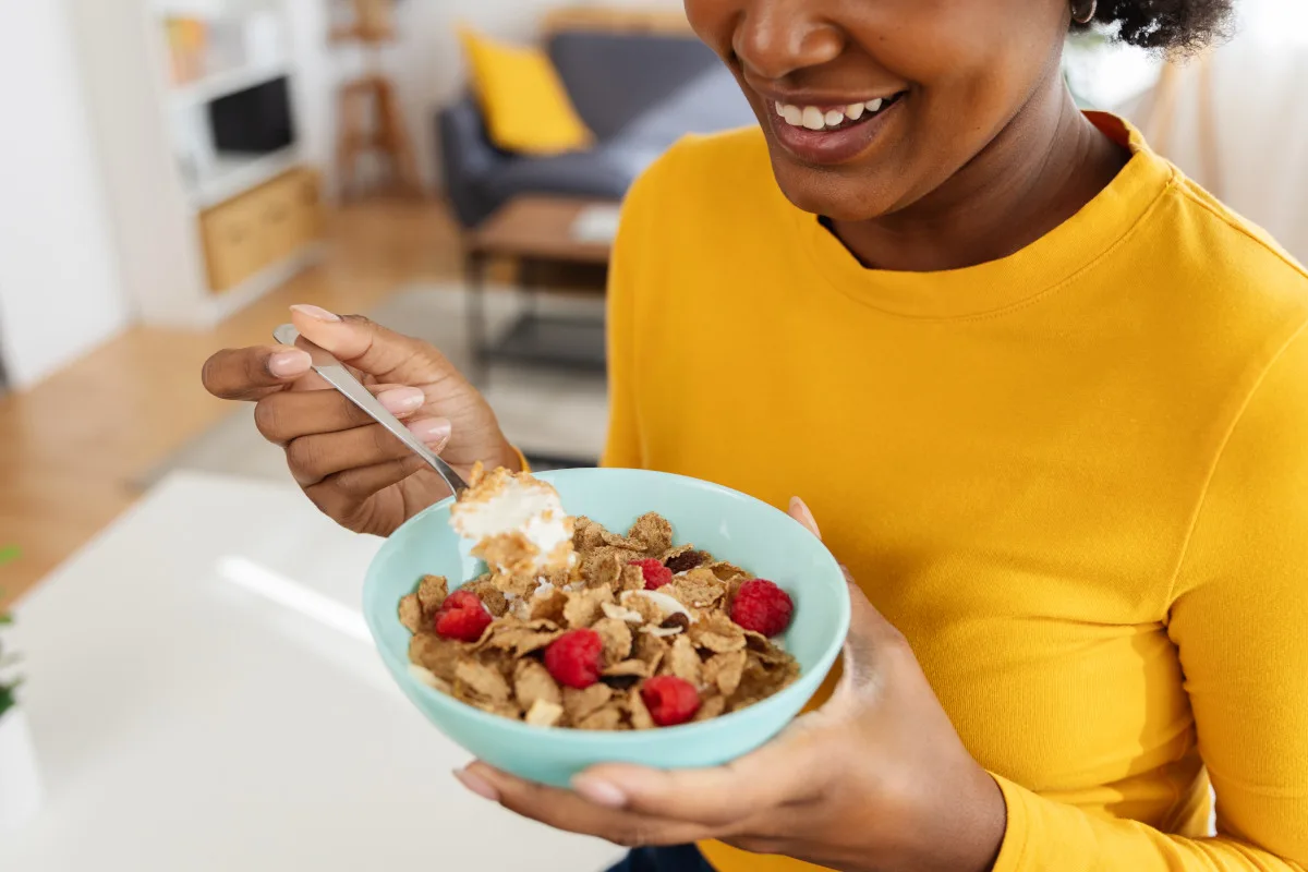 Smiling woman enjoying a healthy breakfast of cereal with raspberries and yogurt at modern apartment.