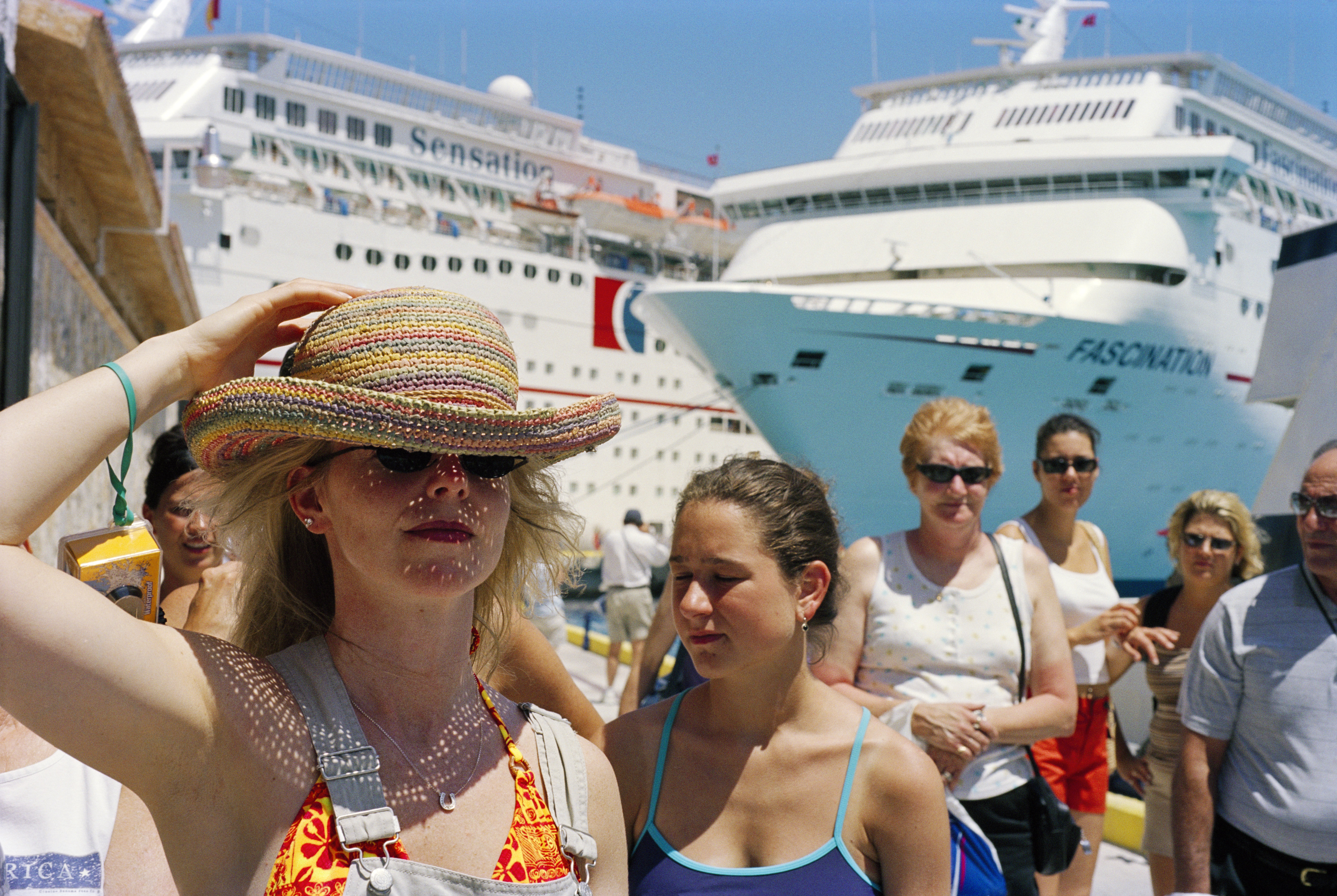 A woman in a sun hat stands in the foreground as two massive white cruise ships, the Sensation and the Fascination, loom in the background at a port.