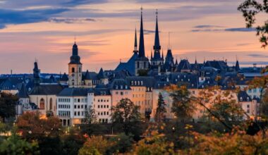 Luxembourg city skyline at sunset with church spires, historic rooftops, and trees displaying autumn foliage under a colourful sky