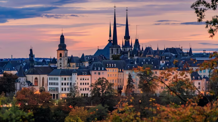 Luxembourg city skyline at sunset with church spires, historic rooftops, and trees displaying autumn foliage under a colourful sky