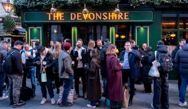A crowd of people stand outside The Devonshire pub, socializing and drinking on the street.