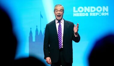 Nigel Farage speaks on stage with his hand raised in front of a blue backdrop reading ‘London Needs Reform’.