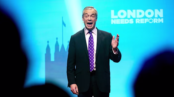 Nigel Farage speaks on stage with his hand raised in front of a blue backdrop reading ‘London Needs Reform’.