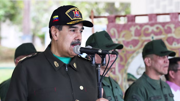 Nicolas Maduro, wearing a military uniform and cap, speaks into a microphone during an event with Venezuelan military personnel.