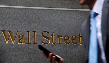 Gold lettering spelling ‘Wall Street’ on a stone wall, with a person in a suit and tie holding a phone in the foreground.