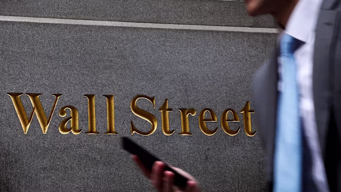 Gold lettering spelling ‘Wall Street’ on a stone wall, with a person in a suit and tie holding a phone in the foreground.