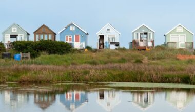 Five colourful beach huts in a row are reflected in still water at sunrise, with long shadows and empty beach grass in the foreground.
