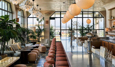 A modern rooftop bar with large windows, globe pendant lights, lush potted plants, and a long marble counter lined with upholstered stools.