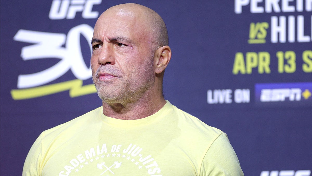 Joe Rogan speaks into a microphone while standing onstage inside an arena.