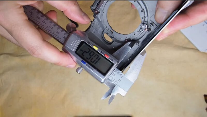 A person uses a digital caliper to measure the width of a camera part. The caliper display reads "29.01 mm". The background is a beige surface.