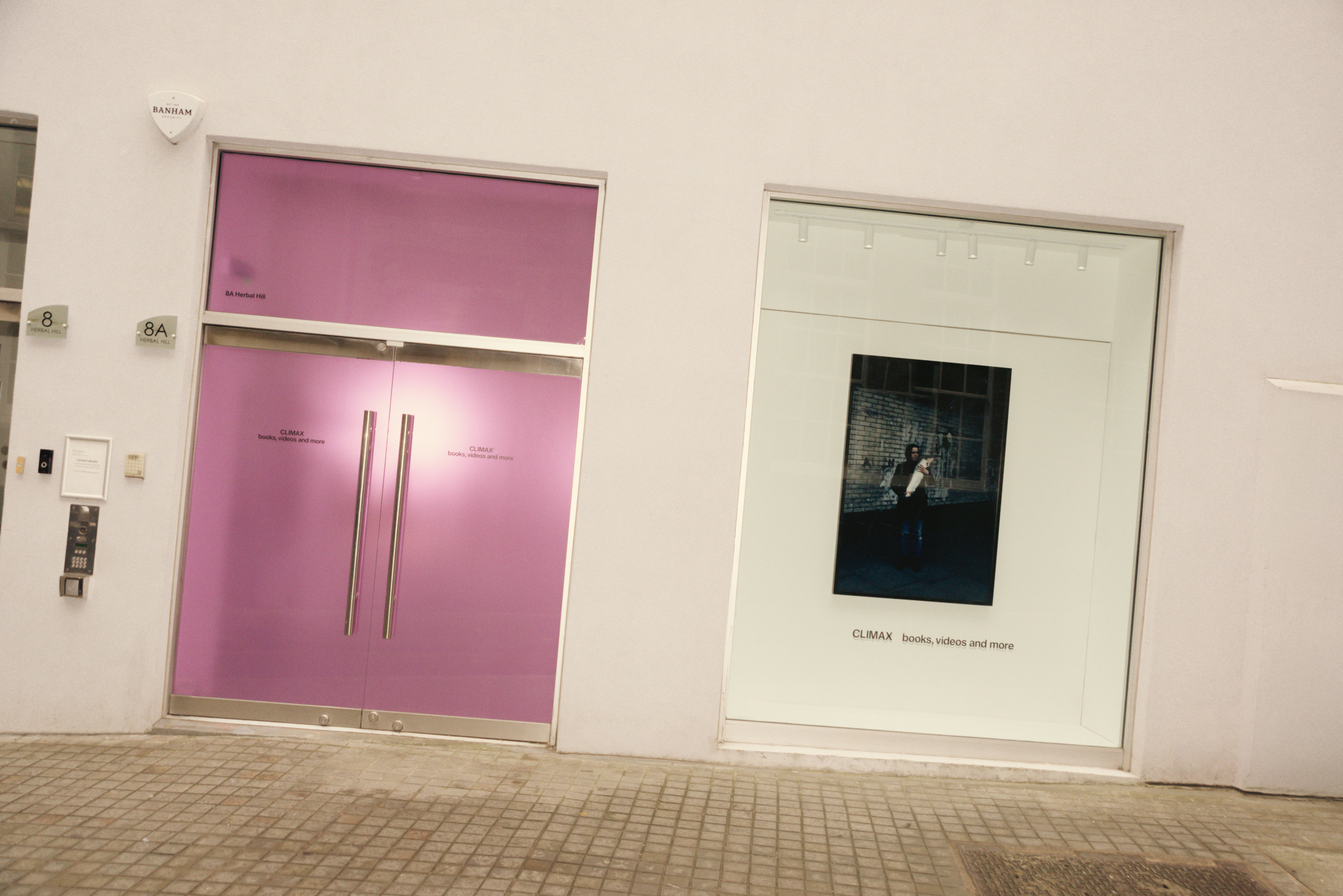 The interiors of a sleekly designed, brightly lit books and merchandise shop features white-washed walls, chrome shelves stakced with colorful tomes, and a pink-hued, see-through door.