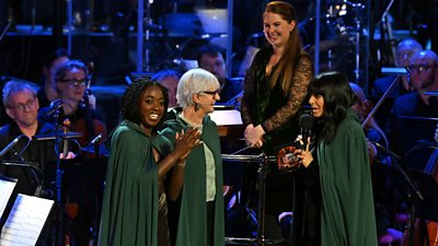 Three women in green cloaks stand in front of an orchestra, smiling and chatting