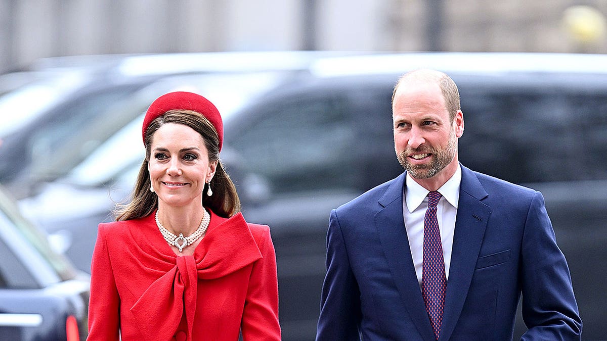 Kate Middleton wearing a red dress and matching hat with pearls walking next to Prince William wearing a blue suit and purple tie.