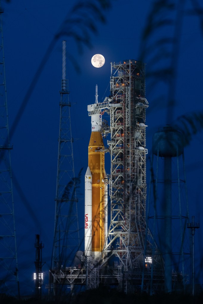 NASA's Space Launch System rocket, which is orange and white, stands on the launch pad with the mobile launcher platform. A full Moon hangs in the sky.