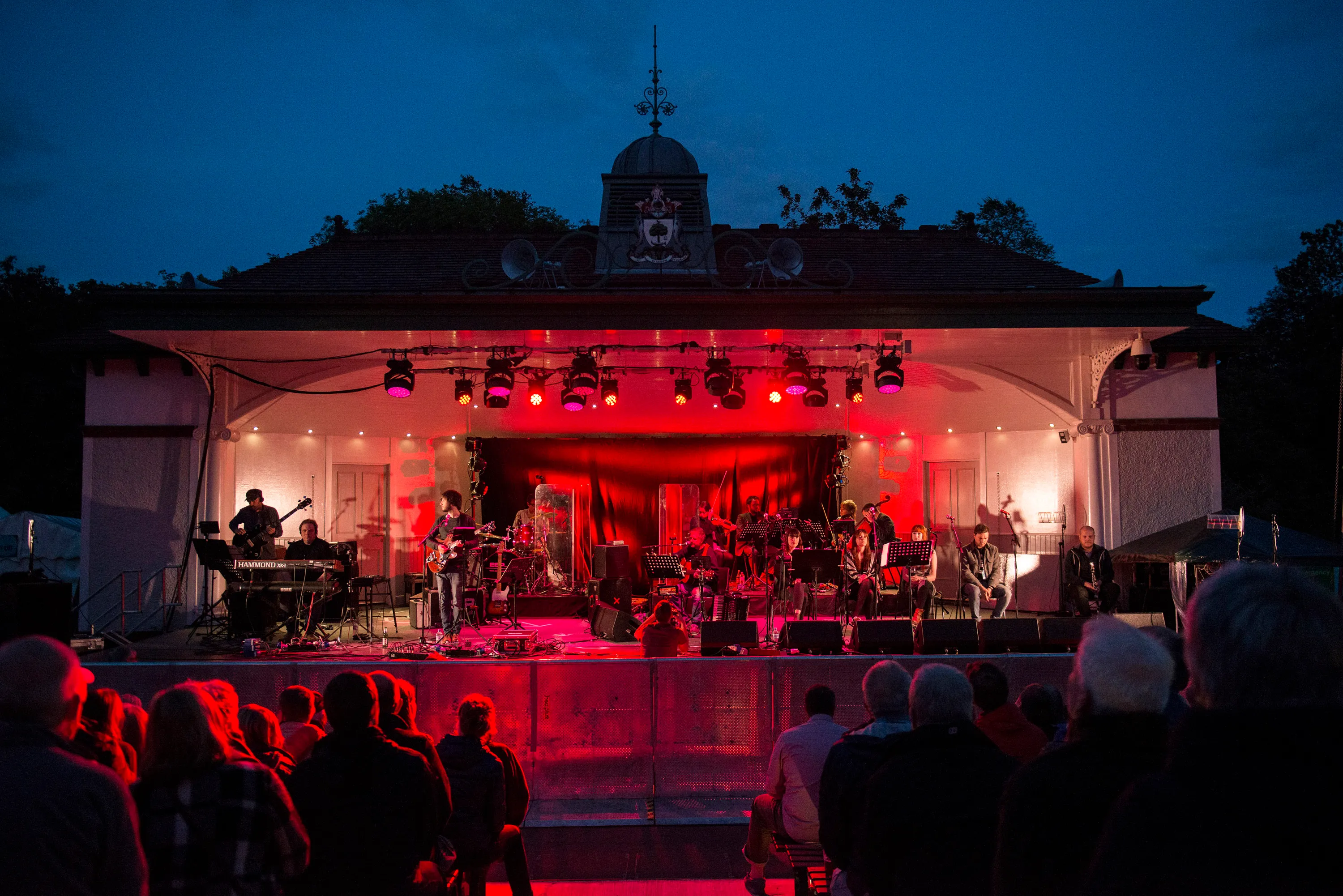 King Creosote performing with a large band on an outdoor stage lit with red lights at night.