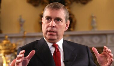 Ex-Prince Andrew wears a suit with a white shirt and a red tie and holds his hands in the air