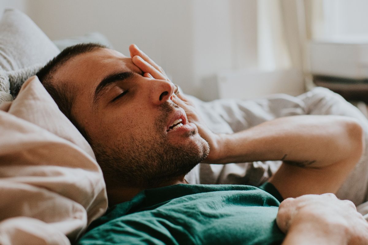 A man has just woken up in a domestic environment, either a living room or a bedroom. He is cozy in duvets and pillows. He rubs his eyes and face sleepily as he comes to.