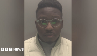 The mugshot of a man wearing transparent glasses and white jacket over a grey jumper.