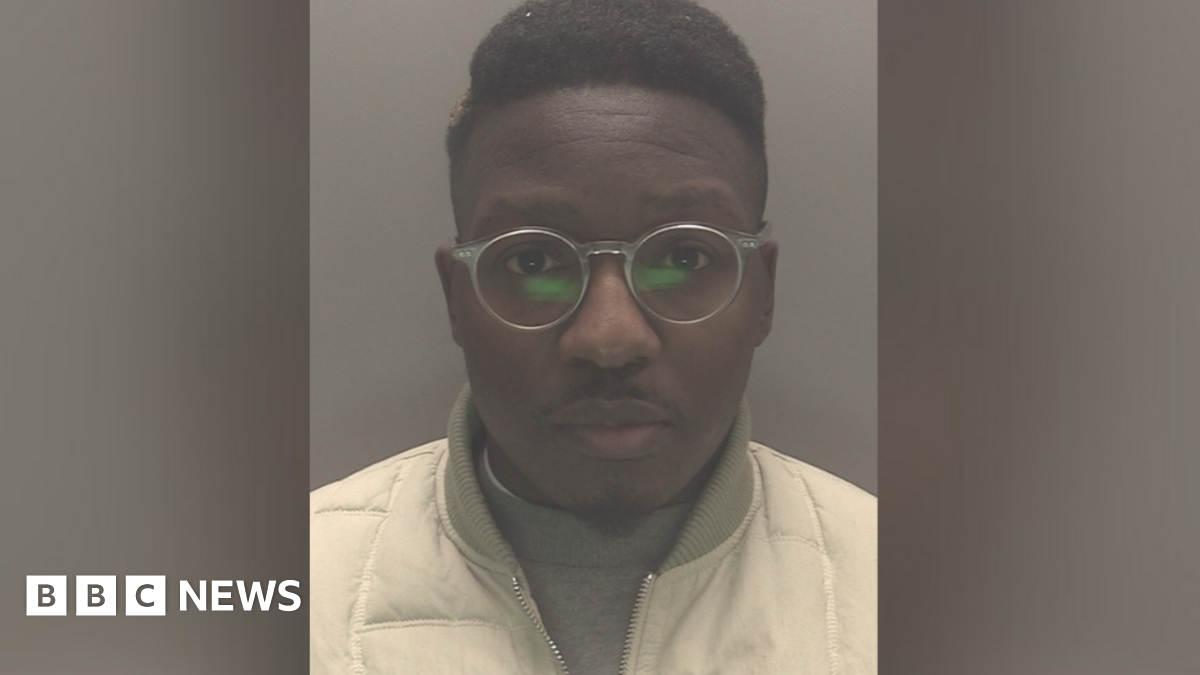 The mugshot of a man wearing transparent glasses and white jacket over a grey jumper.