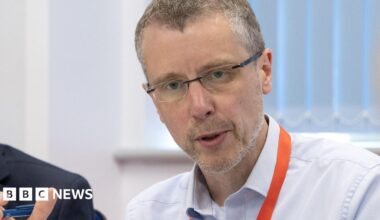Peak Cluster Chief Executive John Egan, who has short grey hair, rimless glasses and who is wearing a light blue shirt with an orange lanyard, pictured mid sentence sat at a table next to a man in a blue suit jacket.