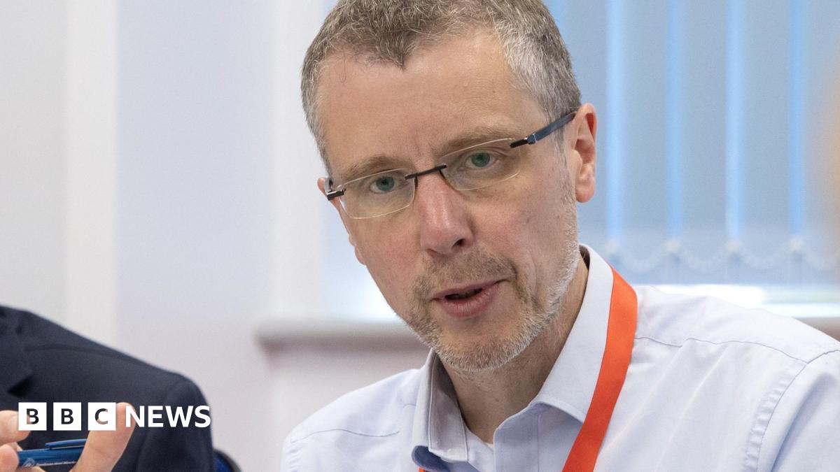 Peak Cluster Chief Executive John Egan, who has short grey hair, rimless glasses and who is wearing a light blue shirt with an orange lanyard, pictured mid sentence sat at a table next to a man in a blue suit jacket.