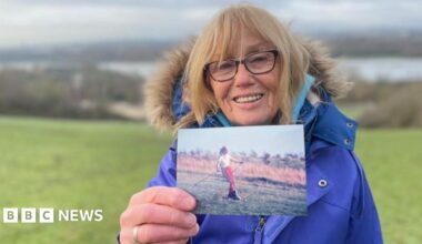 A woman in a thick blue winter jacket and wind-swept blonde hair holds up a picture of a younger woman skiing on grass.