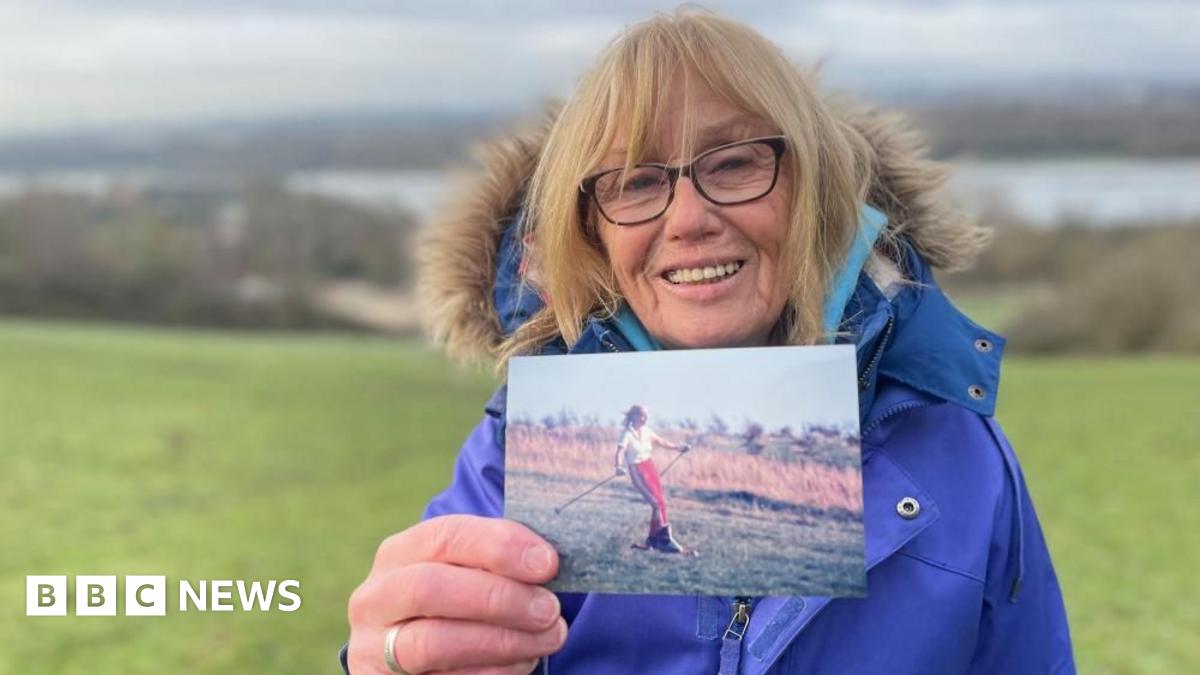A woman in a thick blue winter jacket and wind-swept blonde hair holds up a picture of a younger woman skiing on grass.
