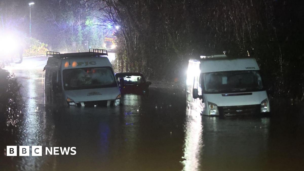 Two white vans and a dark coloured car in between them sit in deep water almost up to the bonnets. Bright lights from emergency vehicles shine in the background in the darkness