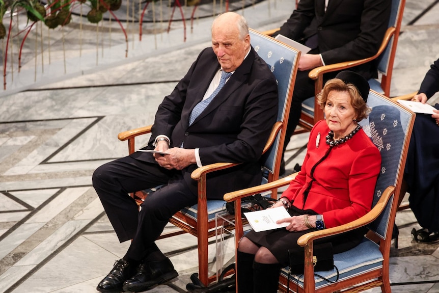 A man and a woman in formal attire, sitting on chairs and looking on.