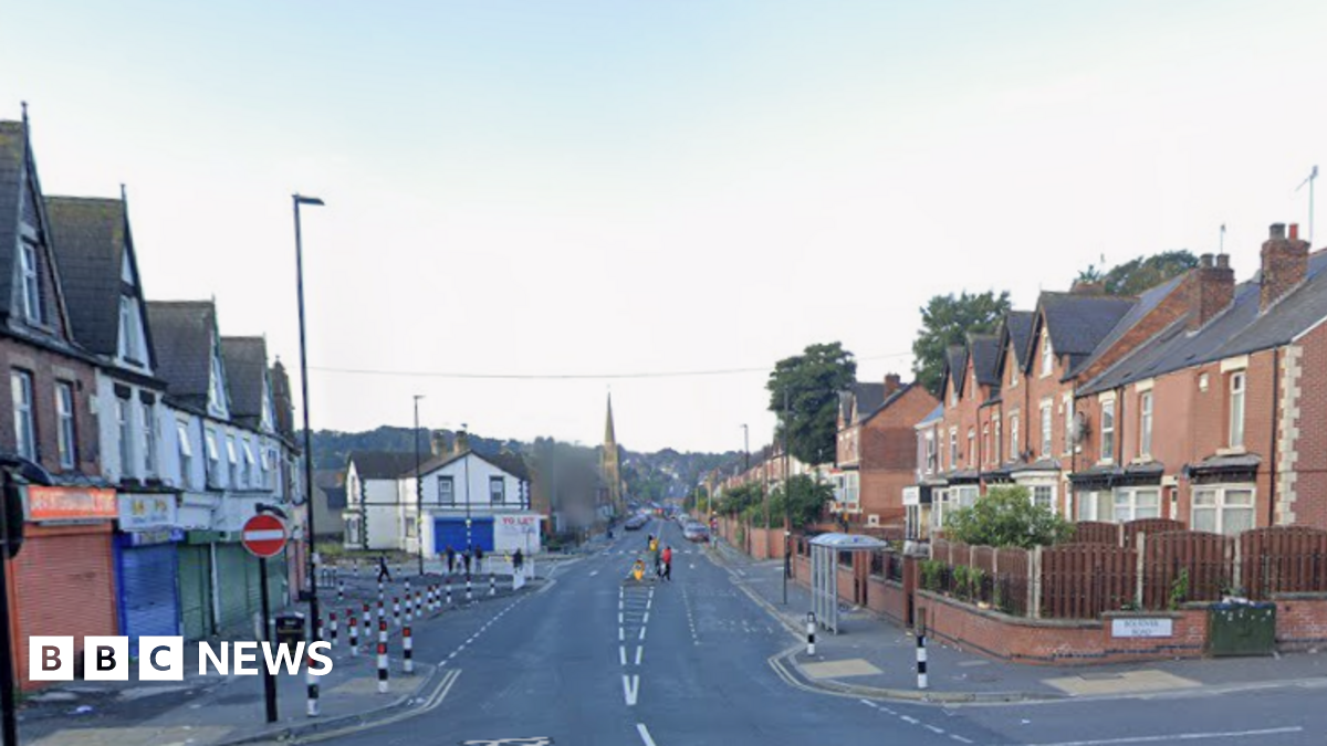 An empty suburban street with small shops with shutters down to the left and terraced homes to the right.