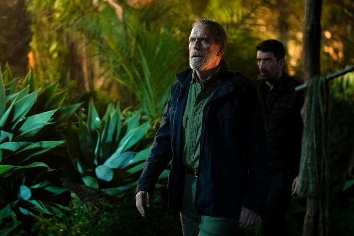 Hugh Laurie stars in The Night Manager season 2; in this scene, Roper is walking in a forest setting, looking dismayed by something