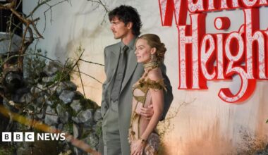 Margot Robbie and Jacob Elordi pose before a moody “Wuthering Heights” backdrop of red lace‑patterned letters, rocks, and branches; one wears a grey suit, the other an ornate gold‑and‑green outfit.