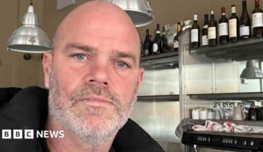 Joel is a bald man with a short grey beard and blue eyes. The selfie is taken in front of metal shelving with bottles of wine on them.