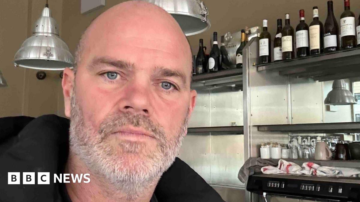 Joel is a bald man with a short grey beard and blue eyes. The selfie is taken in front of metal shelving with bottles of wine on them.