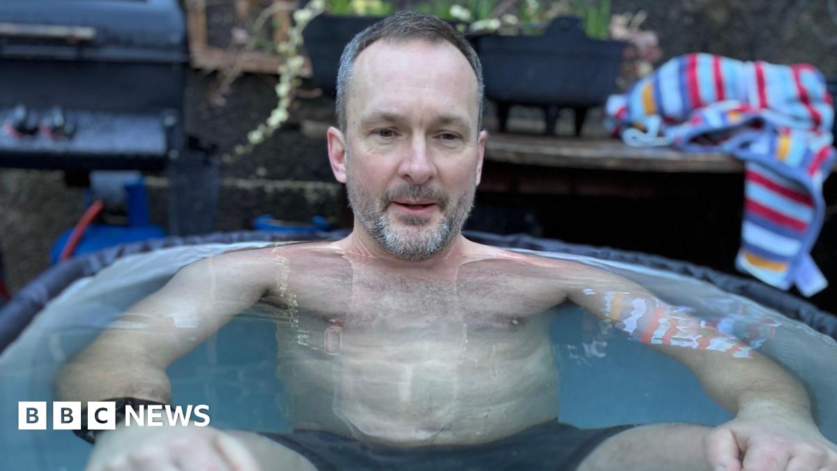 Gerwyn is sitting up to his neck in an ice bath wearing only swimming shorts, looking slightly uncomfortable with a small smile on his face. In the background you can see a blue striped towel and a gas barbecue.