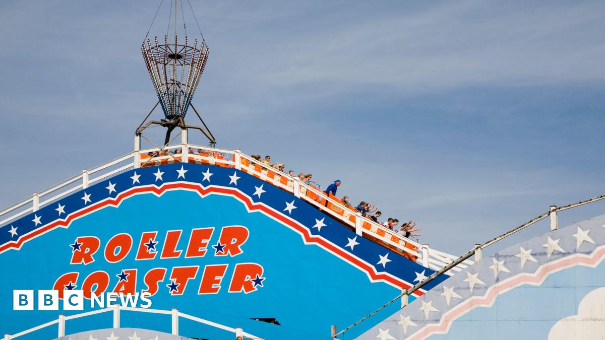 People in carriages heading downwards. Some in the front carriages have their arms in the air. American-style red writing saying "roller coaster" is painted on a sky-blue side of the ride with white stars, and against a navy and red trim bordering the ride. The carriages are orange.