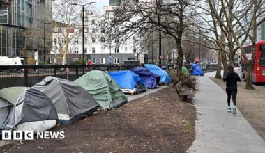 The side of a road is lined with tents and person runs past them on the path. A London red bus is seen going by on the right hand side of the runner.