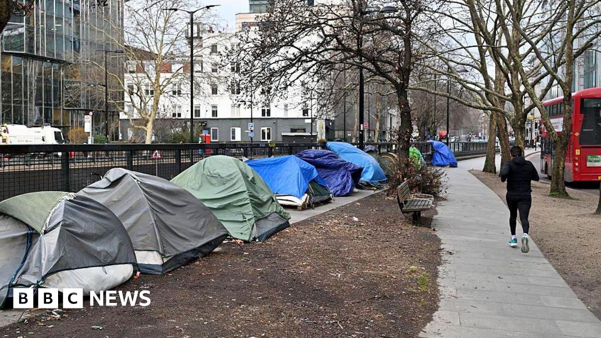 The side of a road is lined with tents and person runs past them on the path. A London red bus is seen going by on the right hand side of the runner.