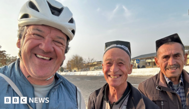 Stephen with two men smiling wearing traditional caps by the side of a road. Stephen is wearing a white cycling helmet and a lightweight blue jacket.