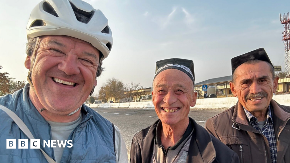 Stephen with two men smiling wearing traditional caps by the side of a road. Stephen is wearing a white cycling helmet and a lightweight blue jacket.