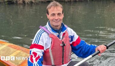 Fourteen people on two boats on a canal in Birmingham. There are 10 women and four men from different ethnicities wearing red life jackets. The boats are parked up by the canal footpath - one at the front is red and the other is purple at the back. There is also a Union Jack flag at the front of the purple boat.