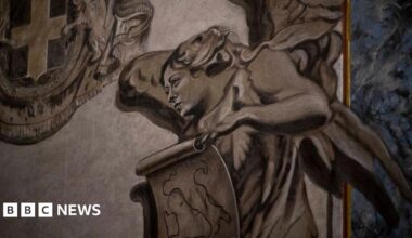 A close-up shot of a painted grey angel. Its face resembles that of Italian Prime Minister Giorgia Meloni. She has wings and holds a scroll on which the shape of Italy is drawn.