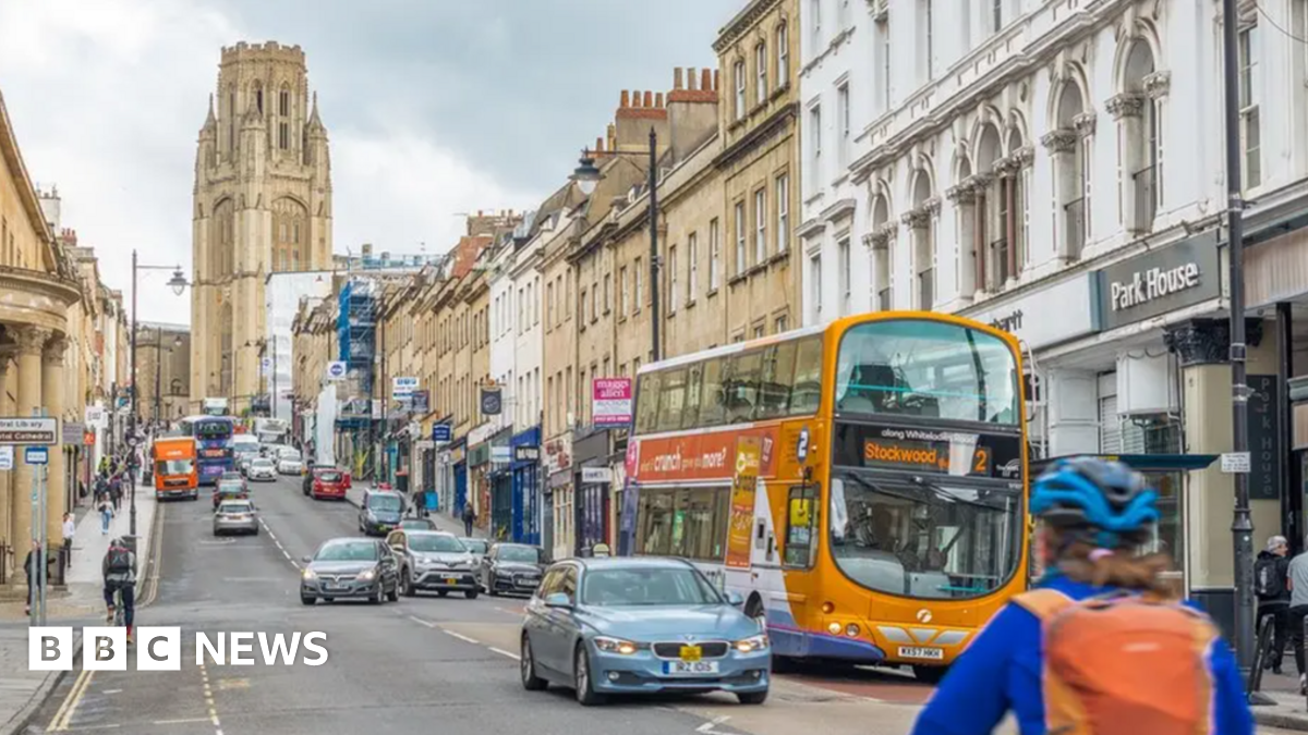 Park Street. It is a busy, hilled road with cars, busses and a cyclist travelling on it. The Wills Memorial building is at the top of it with a grand tower. Shops line the street and pedestrians walk up and down the pavement.