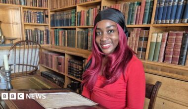 Princess, wearing a black head bandana and red top, sits at a antique table in the library with shelves of books behind her.
