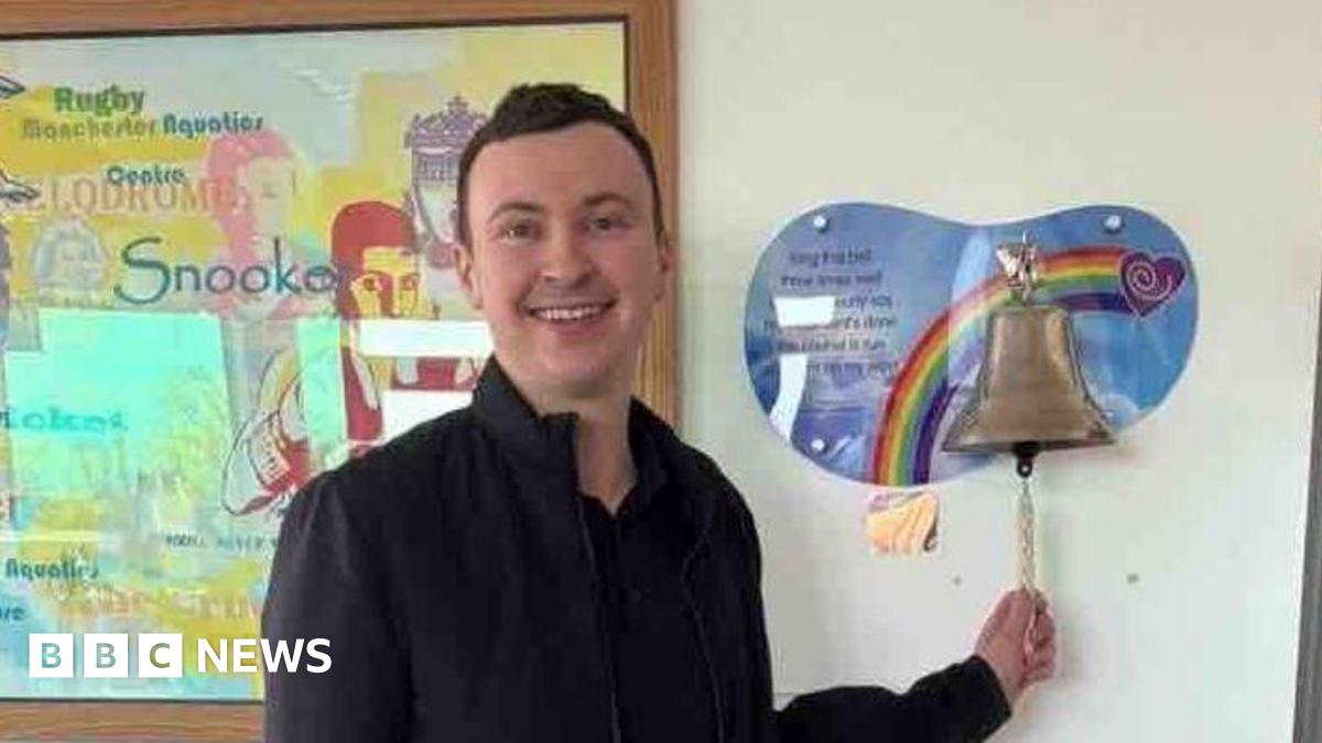 Man with dark brown short hair wears an all black outfit and is smiling - Sam Birchall. The 24-year-old stands in a hospital room in front of a bronze bell on a rainbow mount. He holds the string of the bell as if to begin ringing it.
