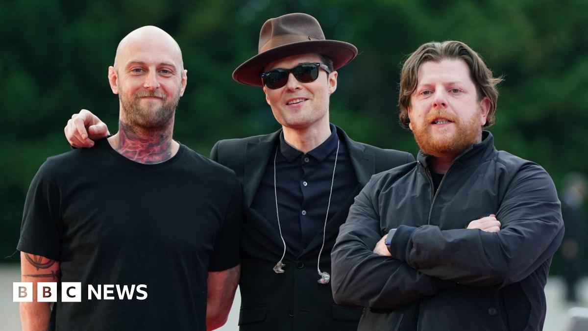Three men stand side by side smiling at the camera. Mince Fratelli, a bald man with a tattooed neck wearing a black t-shirt, is on the far left. Jon Fratelli is in the centre and wears a brown hat, black sunglasses and a dark suit with earphones dangling on his chest. On the right is Barry Fratelli who has brown hair and a ginger beard and wears a dark jacket with his arms crossed across his chest.