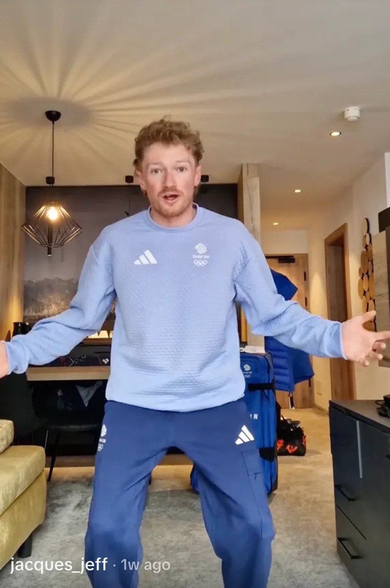 A person wearing a sporty tracksuit poses energetically in a modern living room with open arms