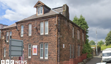A red brick building, with railings around it. It is the last building on the street, with a road running upwards next to it and cars parked beside it.