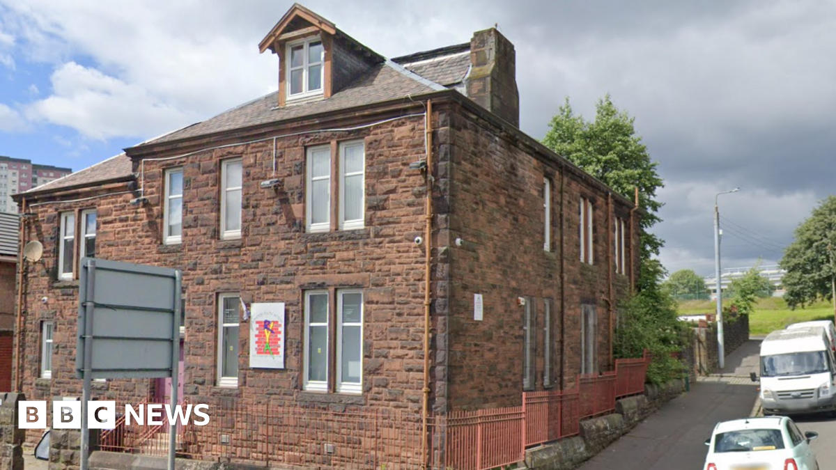 A red brick building, with railings around it. It is the last building on the street, with a road running upwards next to it and cars parked beside it.